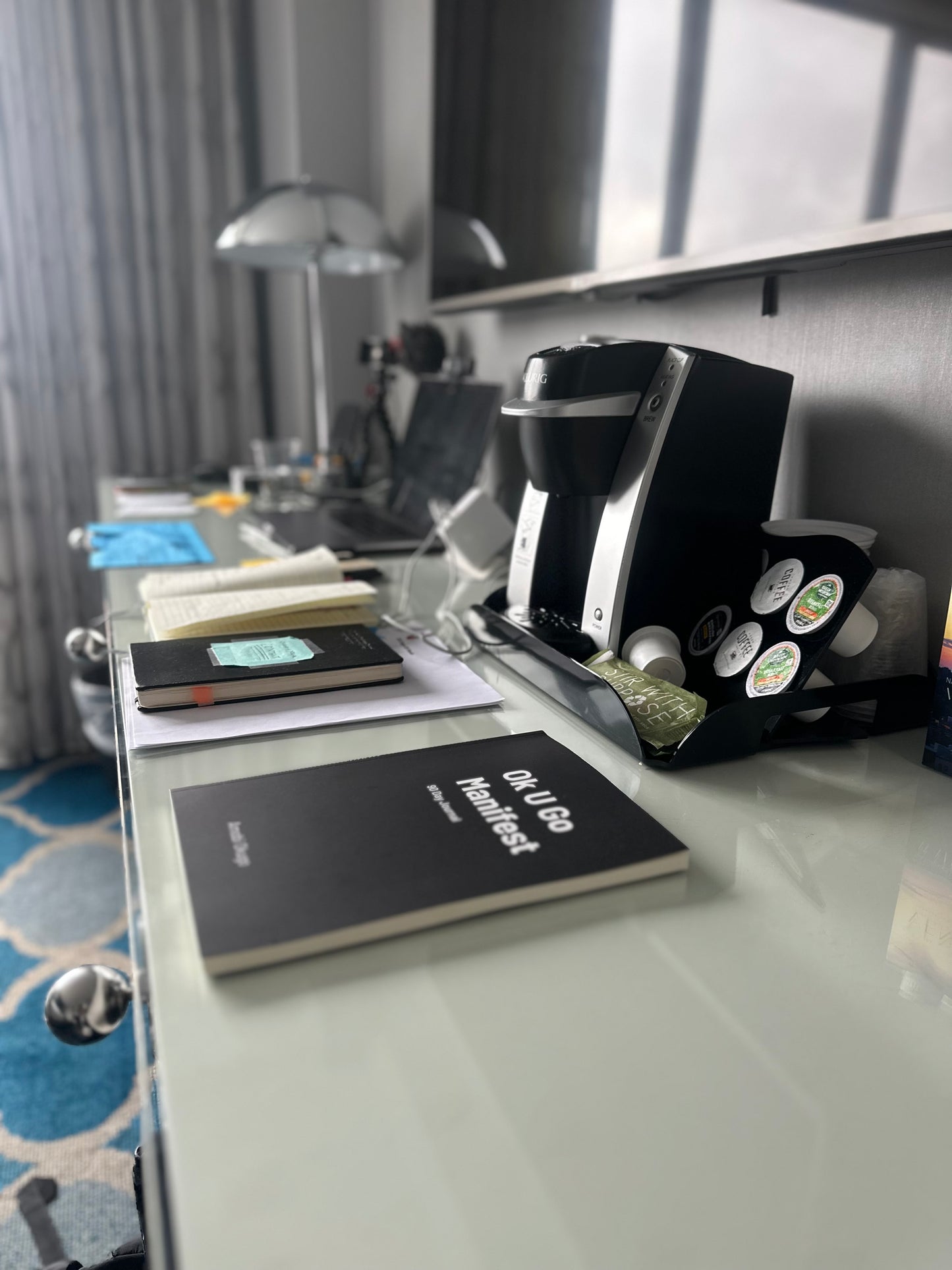 The "Ok U Go Manifest 90 Day Journal" by Amobi Okugo on a hotel desk near a coffee maker and lamp.