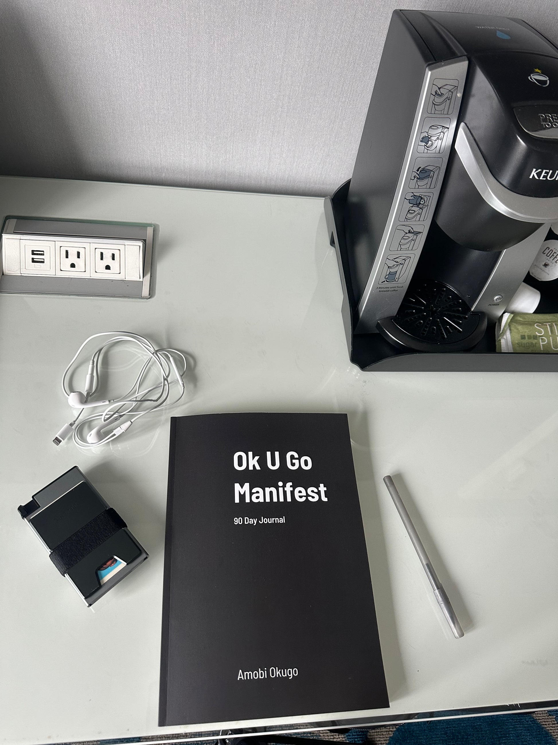 The "Ok U Go Manifest 90 Day Journal" by Amobi Okugo on a desk with a pen, wallet, and headphones.