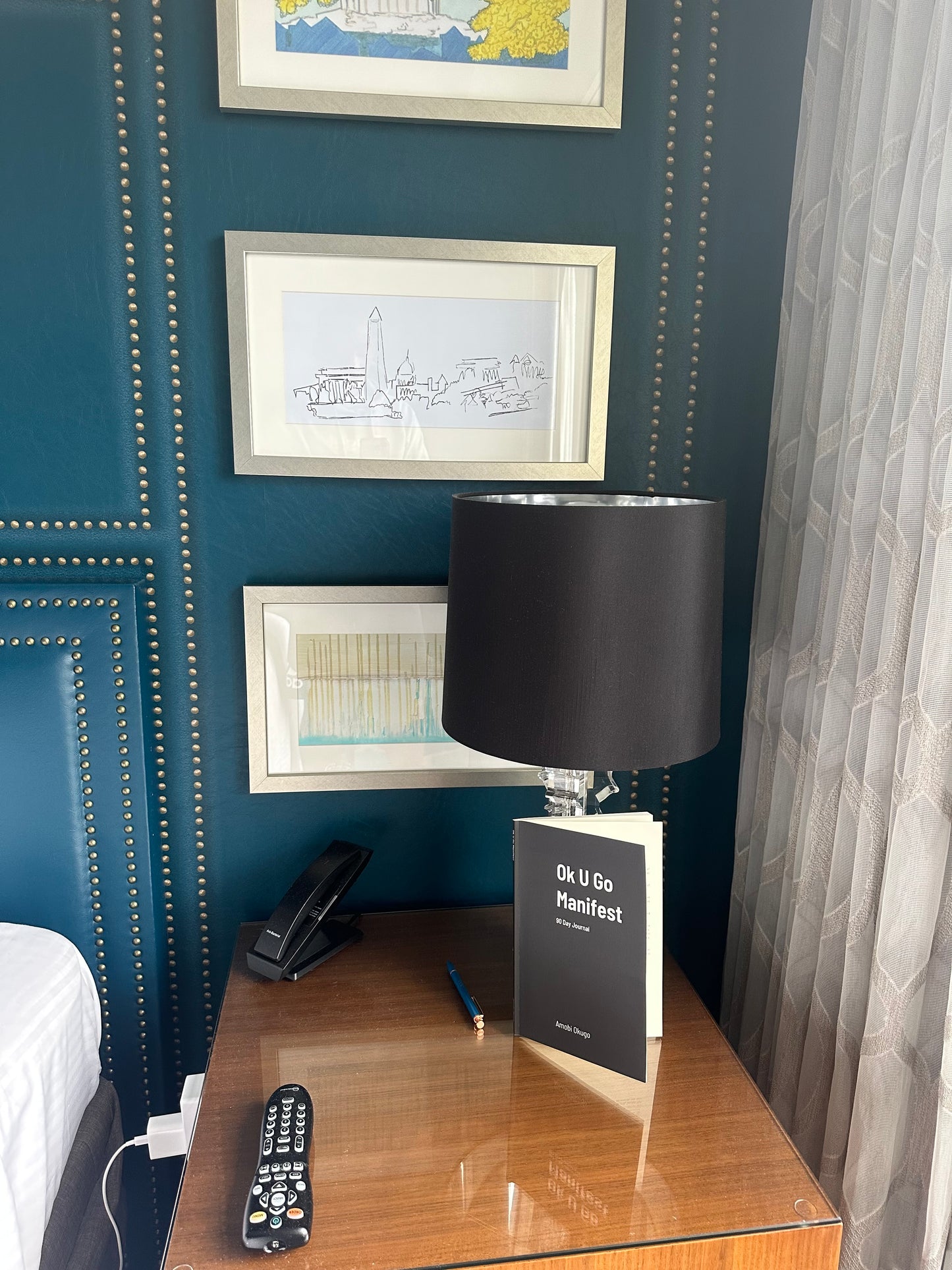 The "Ok U Go Manifest 90 Day Journal" by Amobi Okugo is propped up against a hotel lamp atop a nightstand.